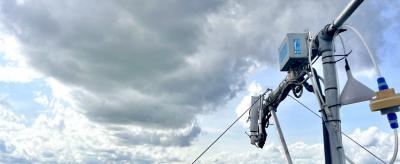 A weather monitoring device is mounted on a pole overlooking a vast forest under cloudy skies.