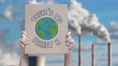 Person holding a sign that reads "there's no planet b" in front of industrial smokestacks emitting smoke against a cloudy sky.