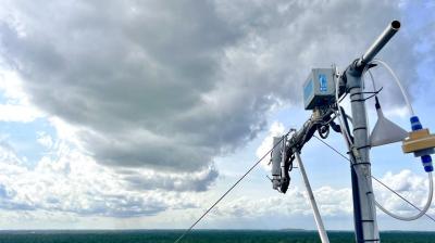 A weather monitoring device is mounted on a pole overlooking a vast forest under cloudy skies.