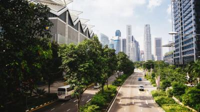 City street with light traffic, lined with trees and modern buildings under a blue sky. Skyscrapers are visible in the background.