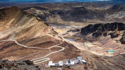 A remote mountainous landscape with winding dirt roads, a cluster of white buildings in the foreground, and rugged, rocky hills and small turquoise lakes in the background.