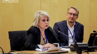 A woman speaks into a microphone at a conference table, while a man sits beside her. Both have conference badges. GEO Group on Earth Observations logo is in the corner.