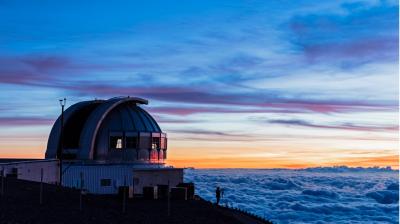 A large observatory dome sits above a sea of clouds at sunset, with a person standing nearby on a mountain slope.