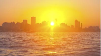 A bright orange sunset over a city skyline, with the sun low above the horizon and water in the foreground.